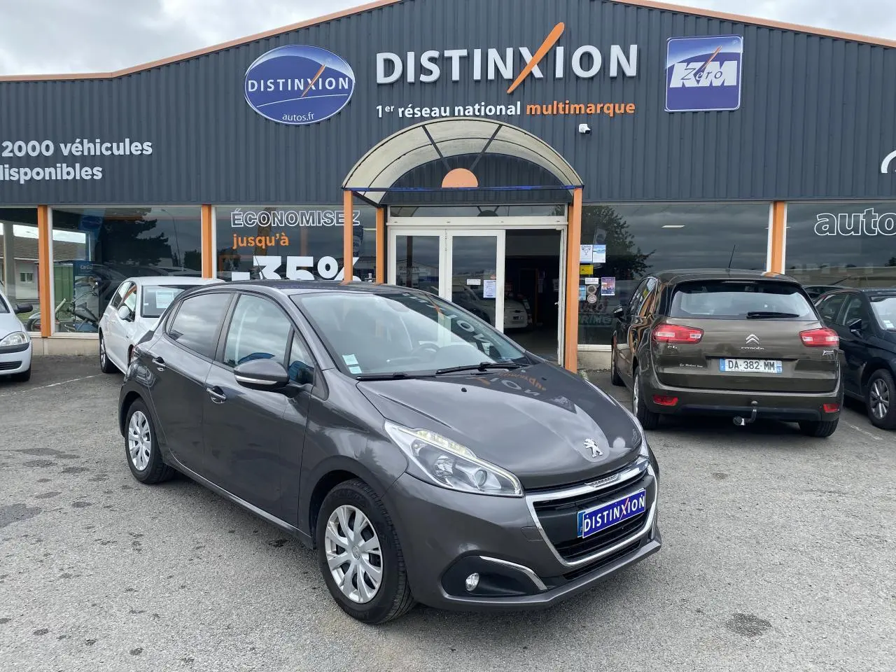 Peugeot 208 gris métallisé vue 3/4 avant droit, stationnée devant un concessionnaire multimarque.