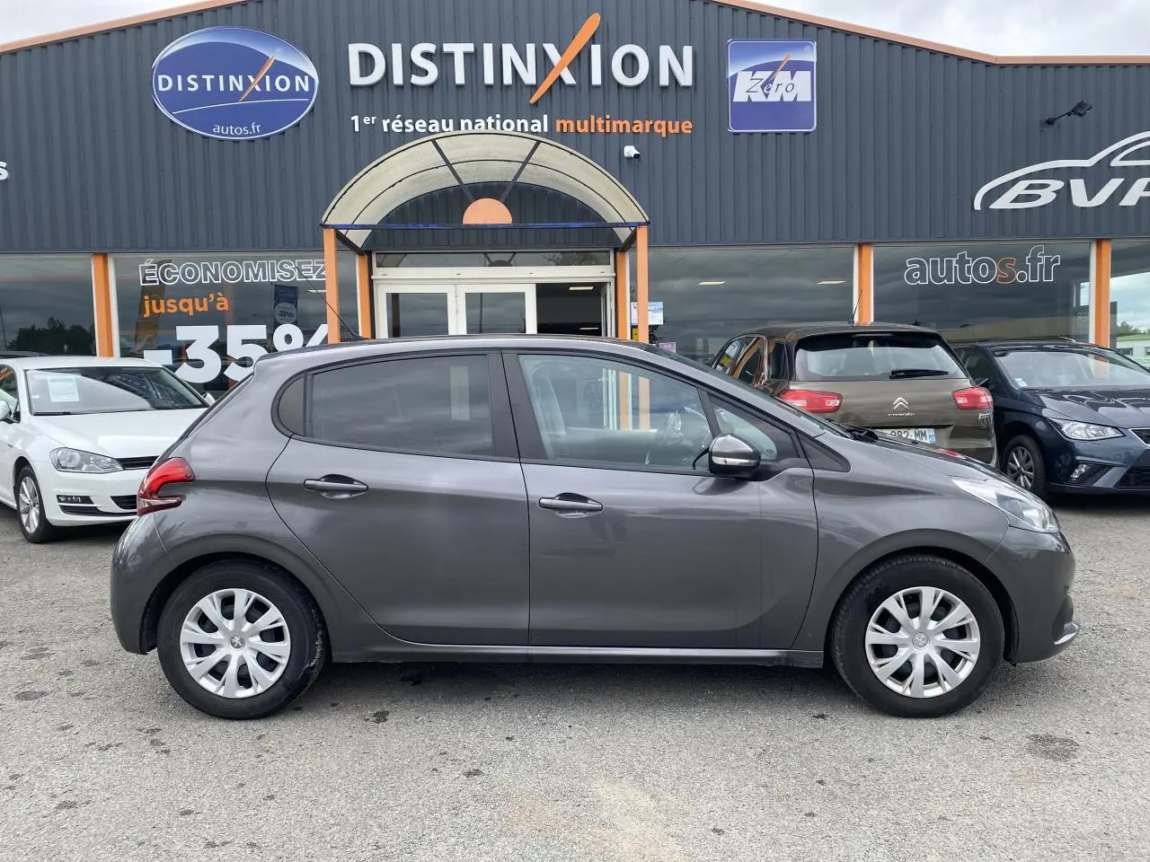 Profil côté gauche d'une Peugeot 208 gris métallisé de 2018, compacte et cinq portes, stationnée devant un concessionnaire.