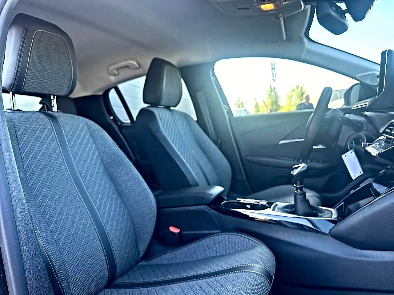 Vue intérieure côté gauche de la Peugeot 208 1.2 100 allure 2025, sièges en tissu gris et boîte manuelle visible.