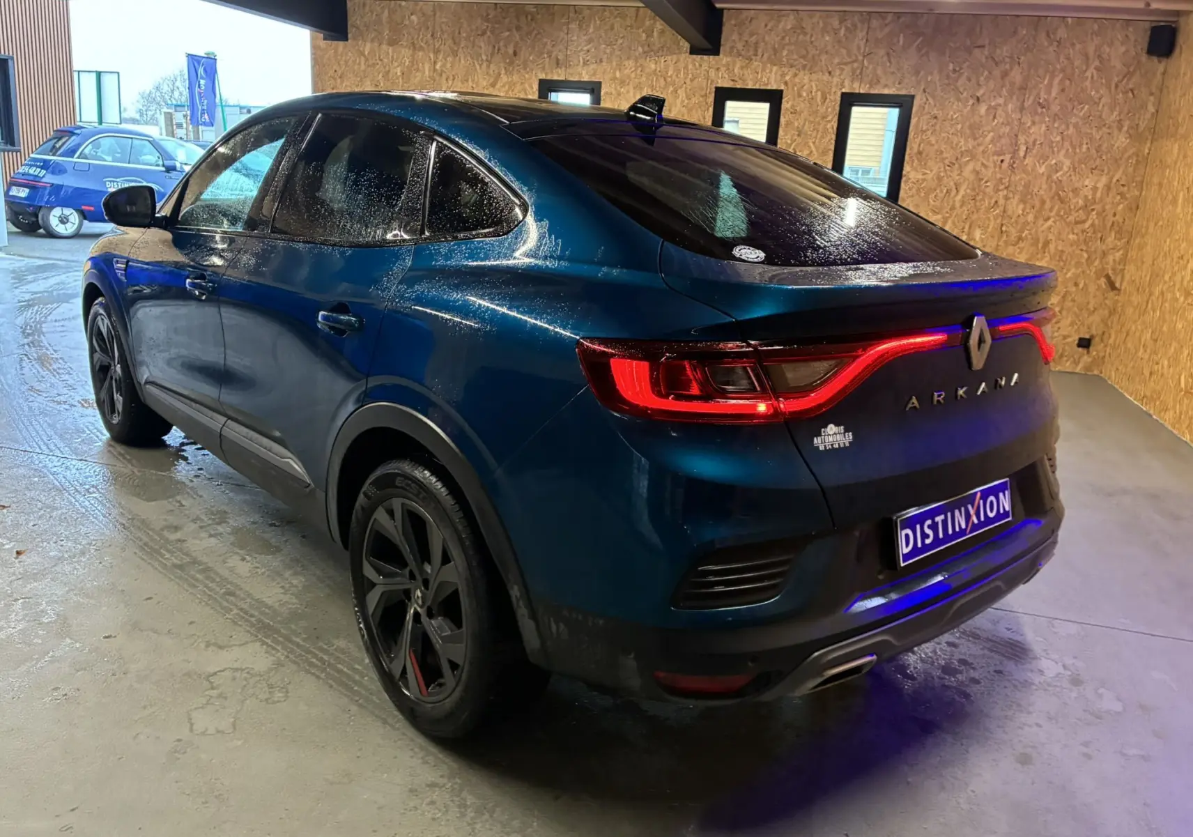 Vue 3/4 arrière droite d’un Renault Arkana bleu 2021 avec feux arrière allumés et jantes noires dans un garage.