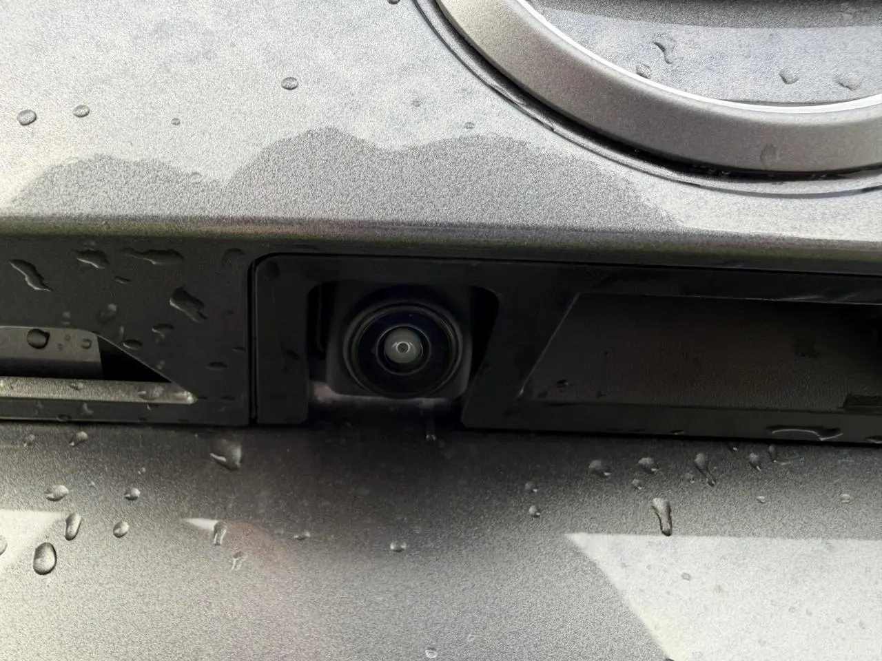 Gros plan sur la caméra de recul intégrée à la plaque arrière du Nissan Juke II noir métal, avec gouttes d'eau visibles.