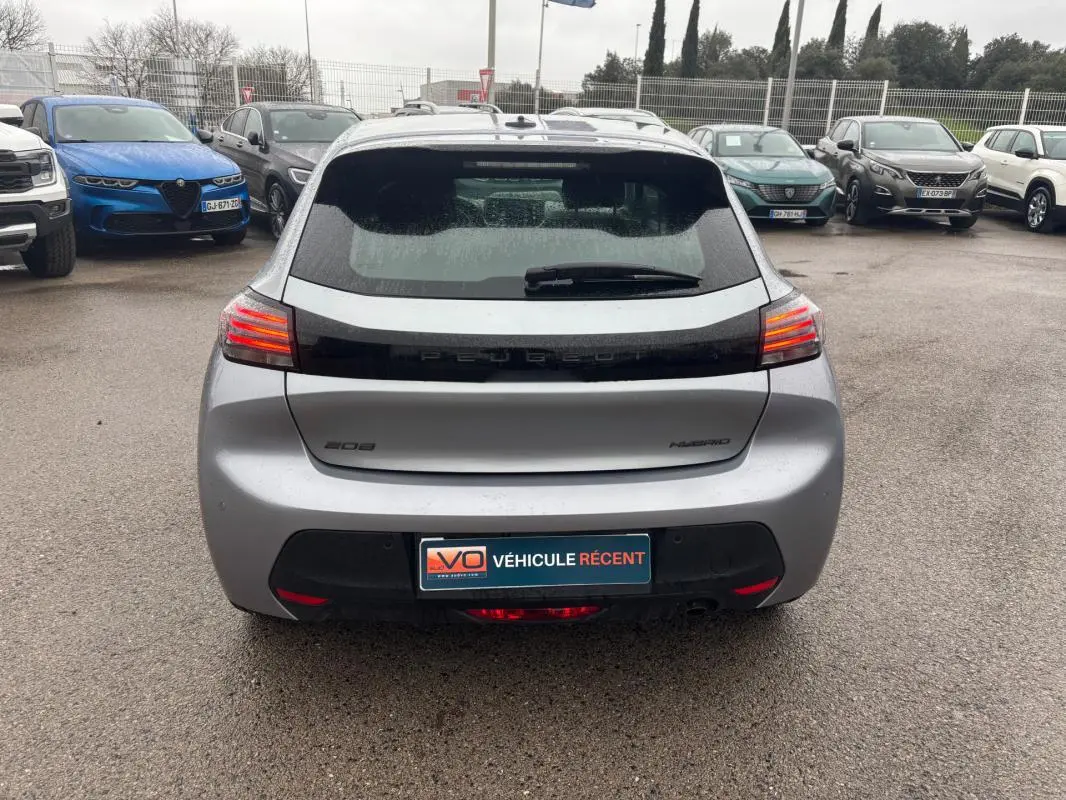 Vue arrière d'une Peugeot 208 Hybrid gris 2025 avec feux arrière allumés sur parking humide.