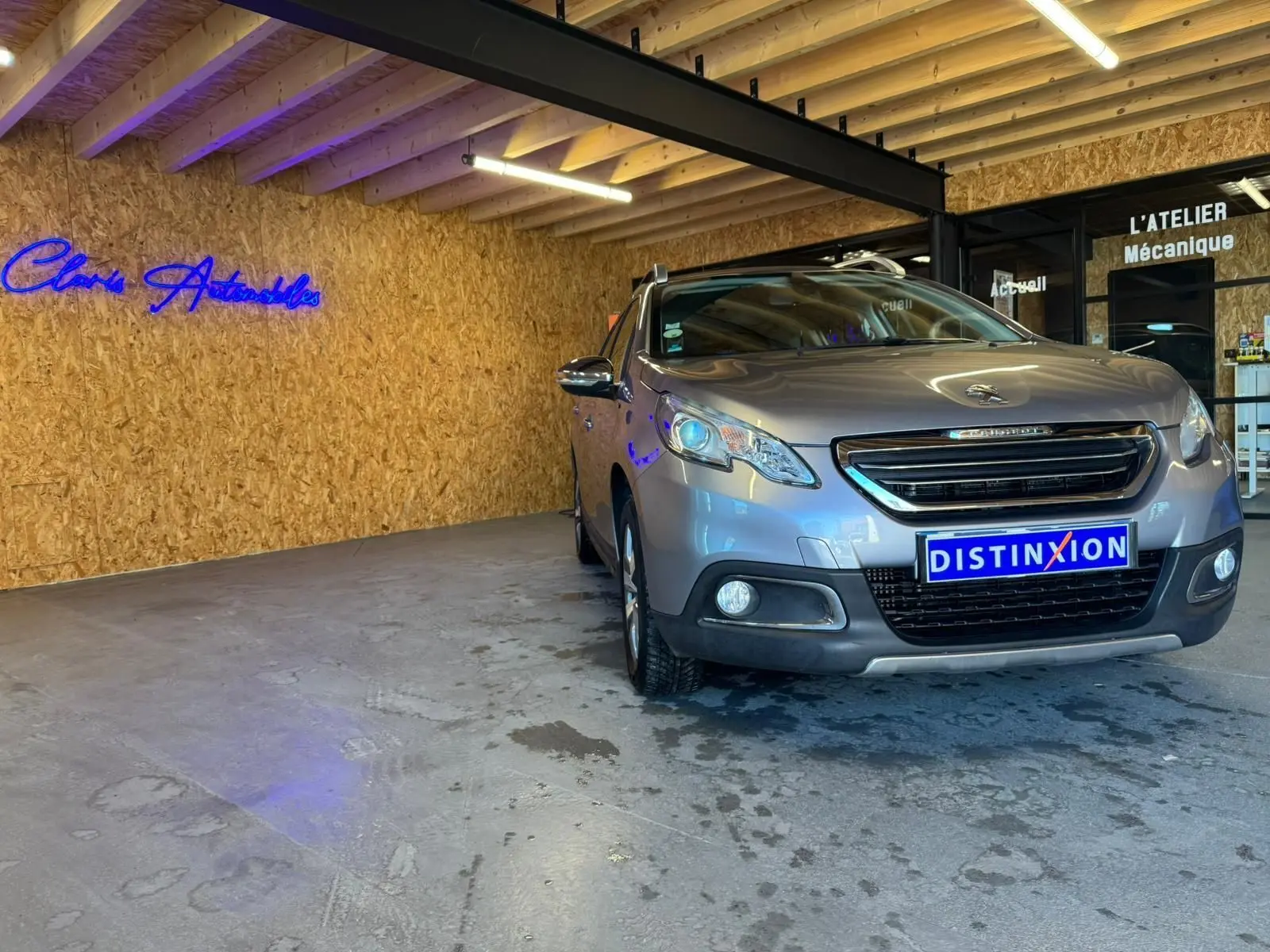 Peugeot 2008 gris en 3/4 avant droit dans un garage avec mur en bois et enseigne lumineuse bleue.