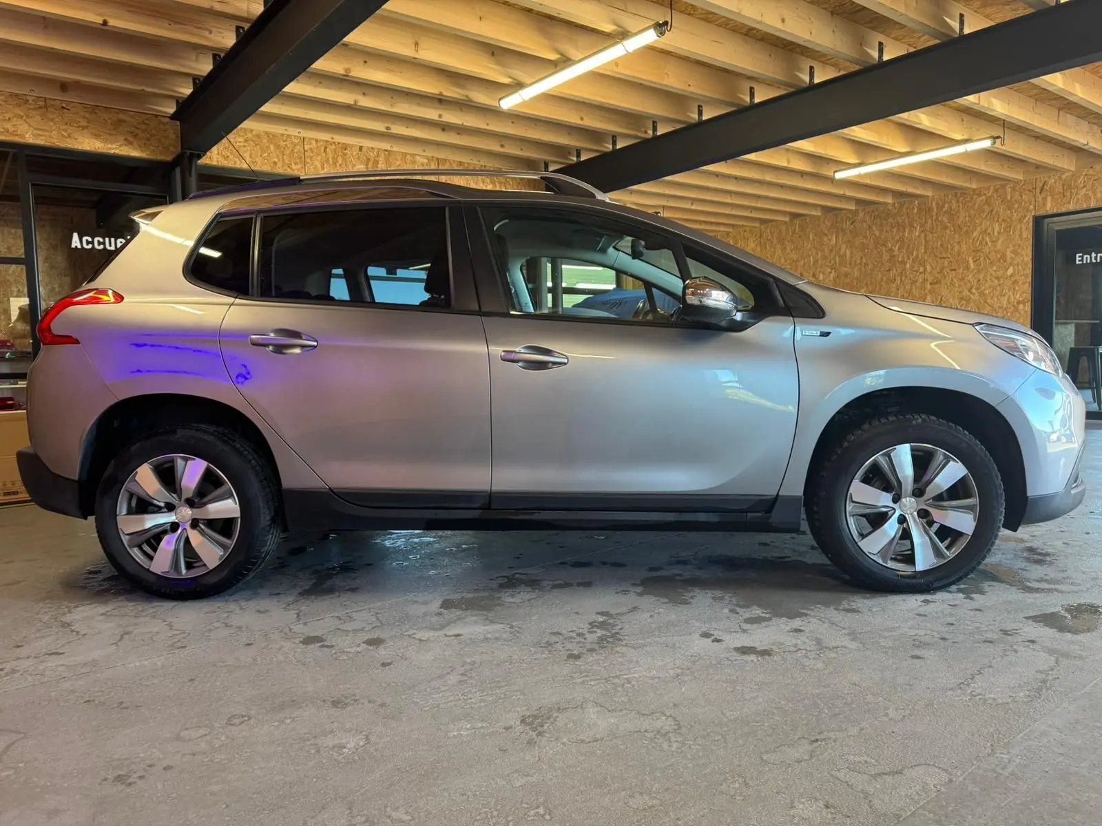 Peugeot 2008 gris vue de profil côté gauche, SUV compact avec jantes alliage et barres de toit en intérieur bois.