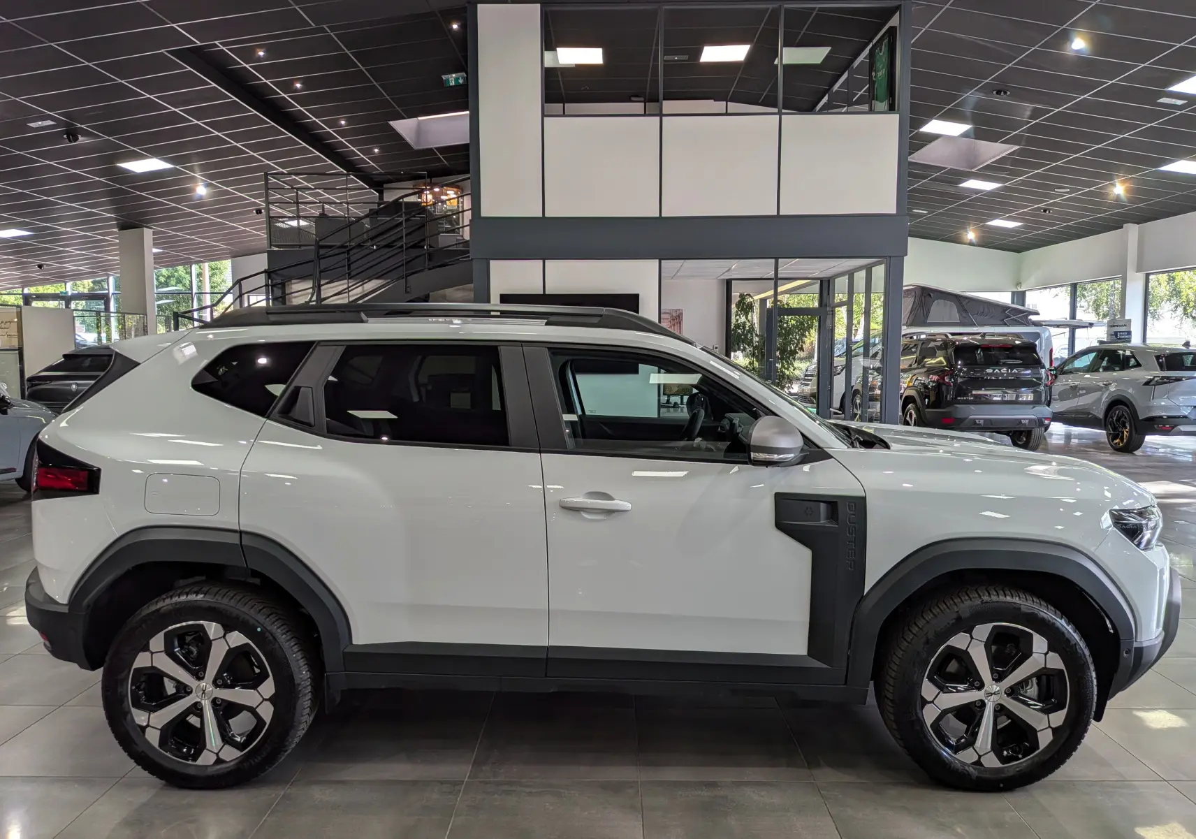 Profil droit d'un Dacia Duster Hybrid-G blanc Glacier en showroom, avec jantes bicolores et protections latérales noires.