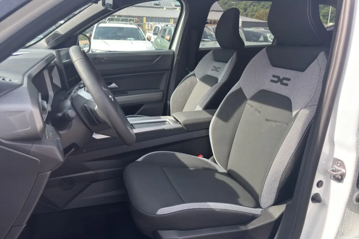 Vue intérieure côté conducteur du Dacia Bigster Hybrid blanc, sièges tissu gris et noir avec logo brodé sur appuie-tête.
