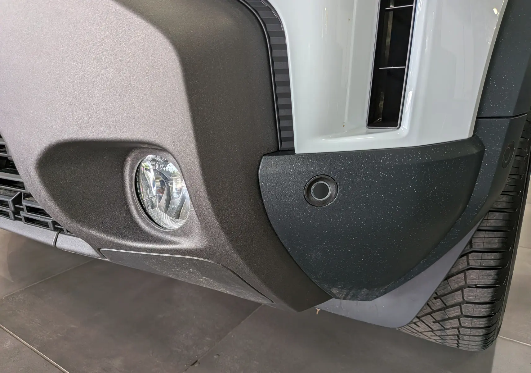 Gros plan sur le pare-chocs avant droit blanc glacier du Dacia Duster Hybrid-G 150 CH 4x4 avec antibrouillard et capteur de stationnement.
