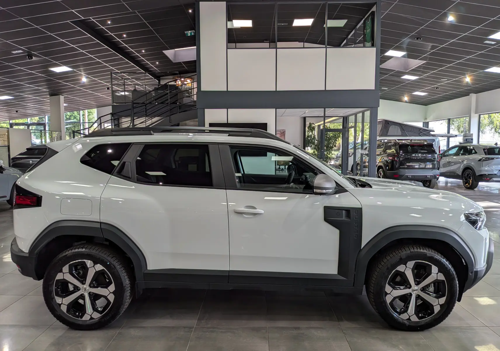 Vue latérale droite d'un Dacia Duster Hybrid-G blanc Glacier 2026 avec jantes bi-ton et protections noires sur les ailes.