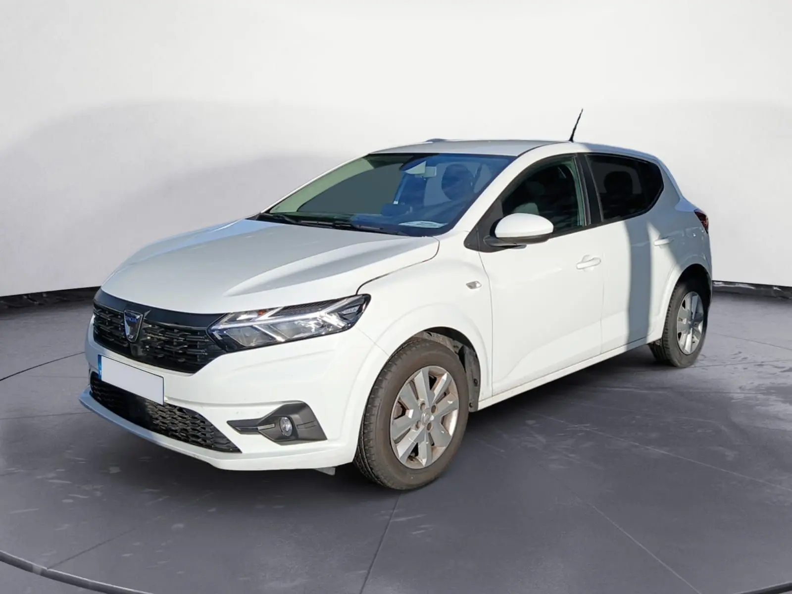 Dacia Sandero blanc vue 3/4 avant droit sur parking gravillonné avec plusieurs véhicules en arrière-plan.