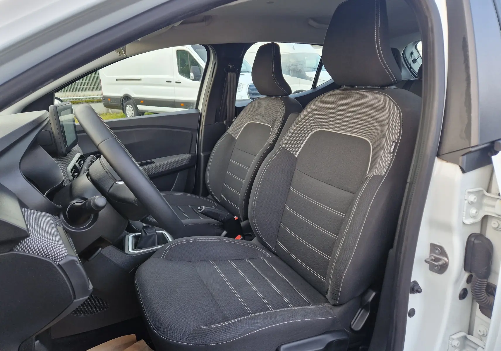 Vue intérieure côté conducteur de la Dacia Sandero blanche 2022, montrant les sièges avant en tissu noir et la console centrale.