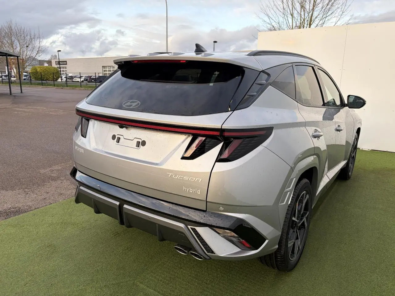 Vue 3/4 arrière droite du Hyundai Tucson 2025 hybride en argent métallisé avec feux arrière à bandeau lumineux et double sortie d'échappement.