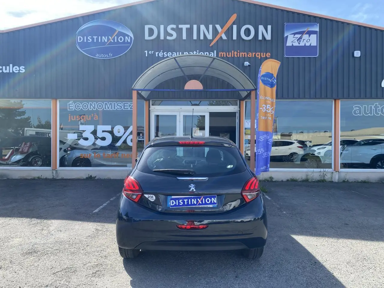Vue arrière d'une Peugeot 208 gris foncé garée devant un showroom Distinxion avec plaques d'immatriculation publicitaires.