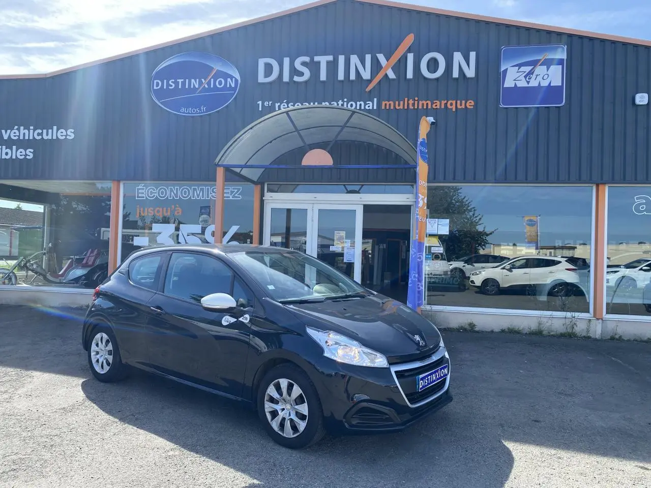 Peugeot 208 gris foncé vue 3/4 avant droit, avec enjoliveurs 15 pouces et rétroviseurs blancs devant un garage Distinxion.