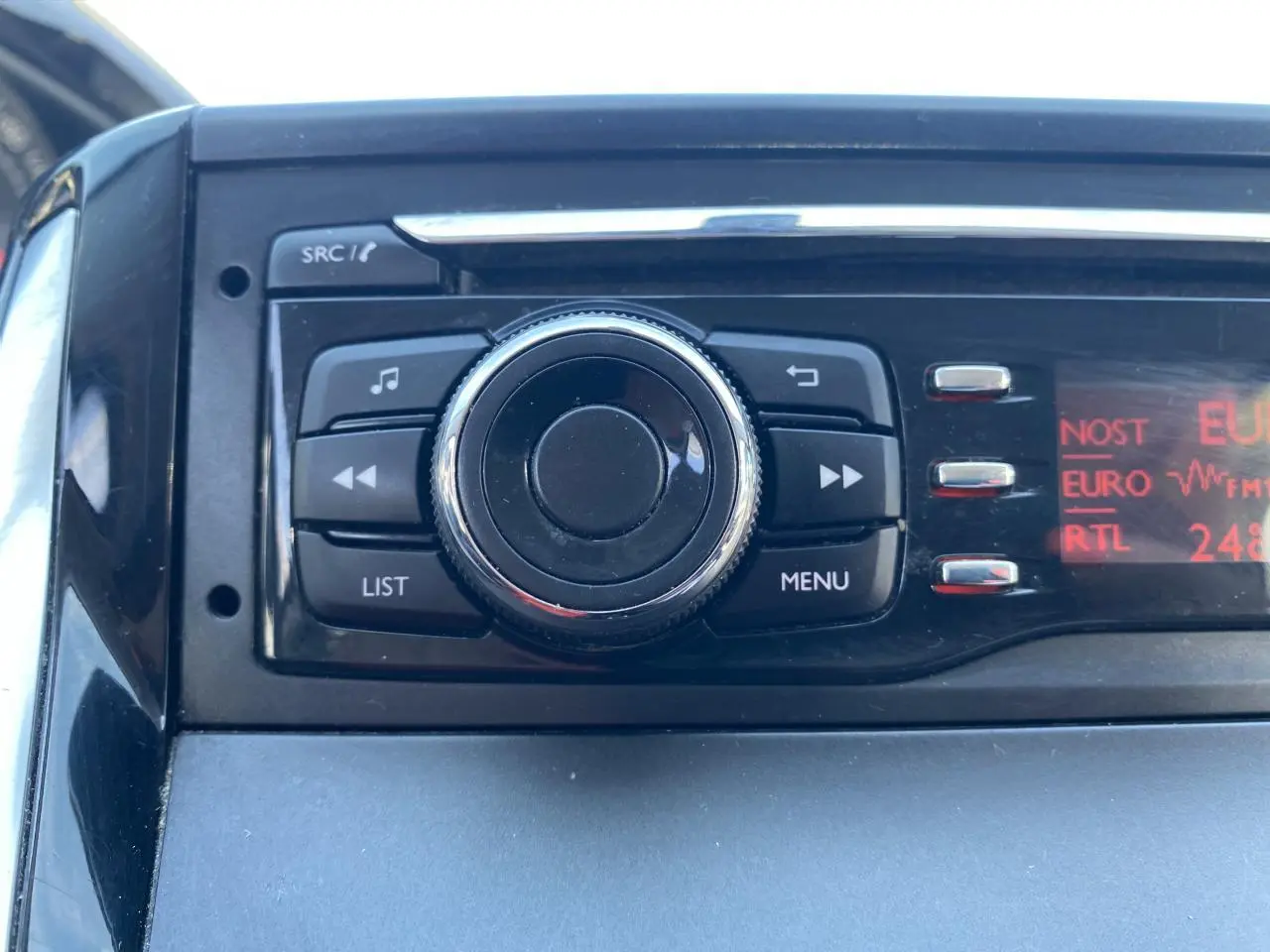 Gros plan sur l'autoradio avec molette centrale et écran rouge affichant les stations, intérieur Peugeot 208 gris foncé.