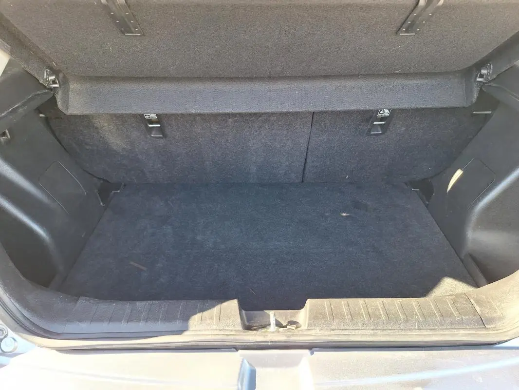 Coffre ouvert d'une Suzuki Baleno gris clair, montrant l'espace de rangement intérieur et la moquette noire.