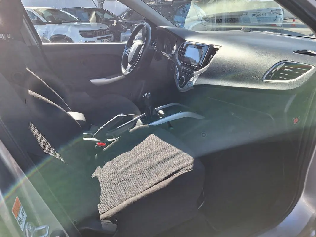Vue intérieure côté conducteur de la Suzuki Baleno gris clair, montrant le volant, la console centrale avec écran tactile et levier de vitesse manuel.