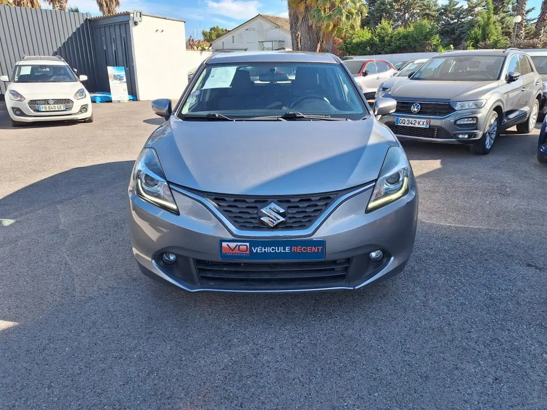 Vue avant d'une Suzuki Baleno gris clair 2017 avec phares allumés et plaque "Véhicule récent" visible.