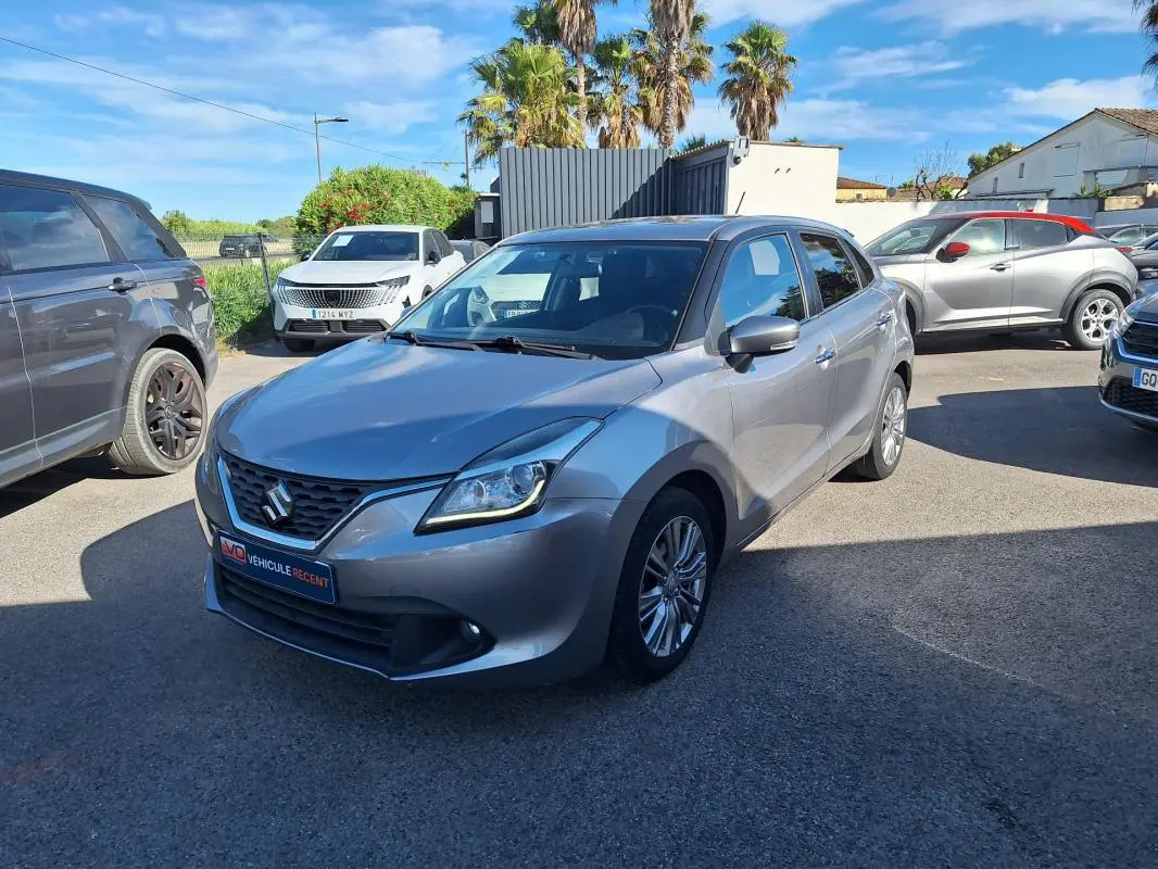 Suzuki Baleno gris clair vue 3/4 avant droit, jantes alliage 16 pouces et phares allumés en extérieur.