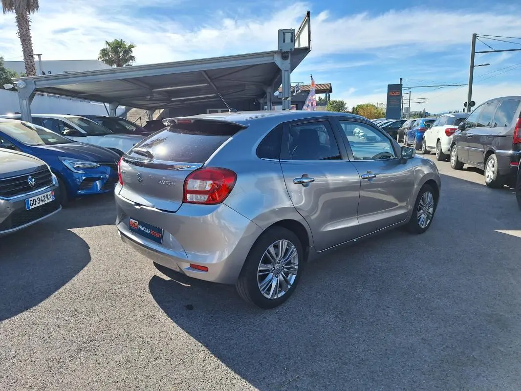 Vue 3/4 arrière droite d'une Suzuki Baleno gris clair avec jantes alliage 16 pouces, vitres surteintées et feux arrière visibles.