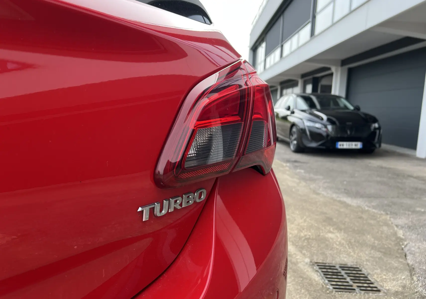Gros plan sur le feu arrière gauche rouge brillant d'une Opel Corsa 1.4 Turbo rouge et noir, avec une Peugeot noire en arrière-plan.