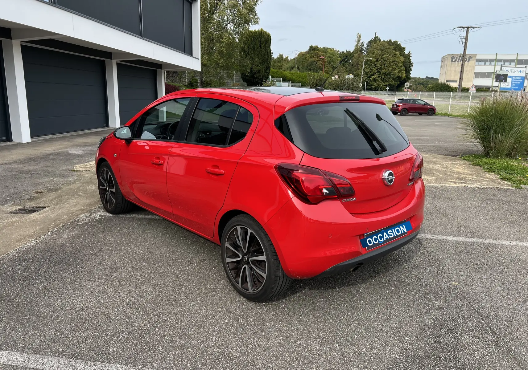 Vue 3/4 arrière droite d'une Opel Corsa rouge avec toit et rétroviseurs noirs brillants sur parking extérieur.