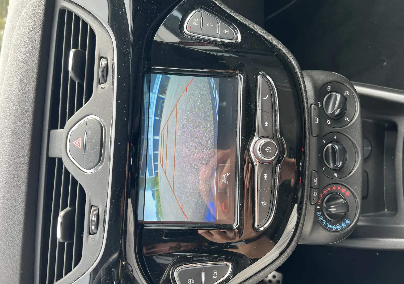 Vue rapprochée de la console centrale noire brillante d'une Opel Corsa 1.4 rouge, avec écran caméra de recul et commandes climatisation.