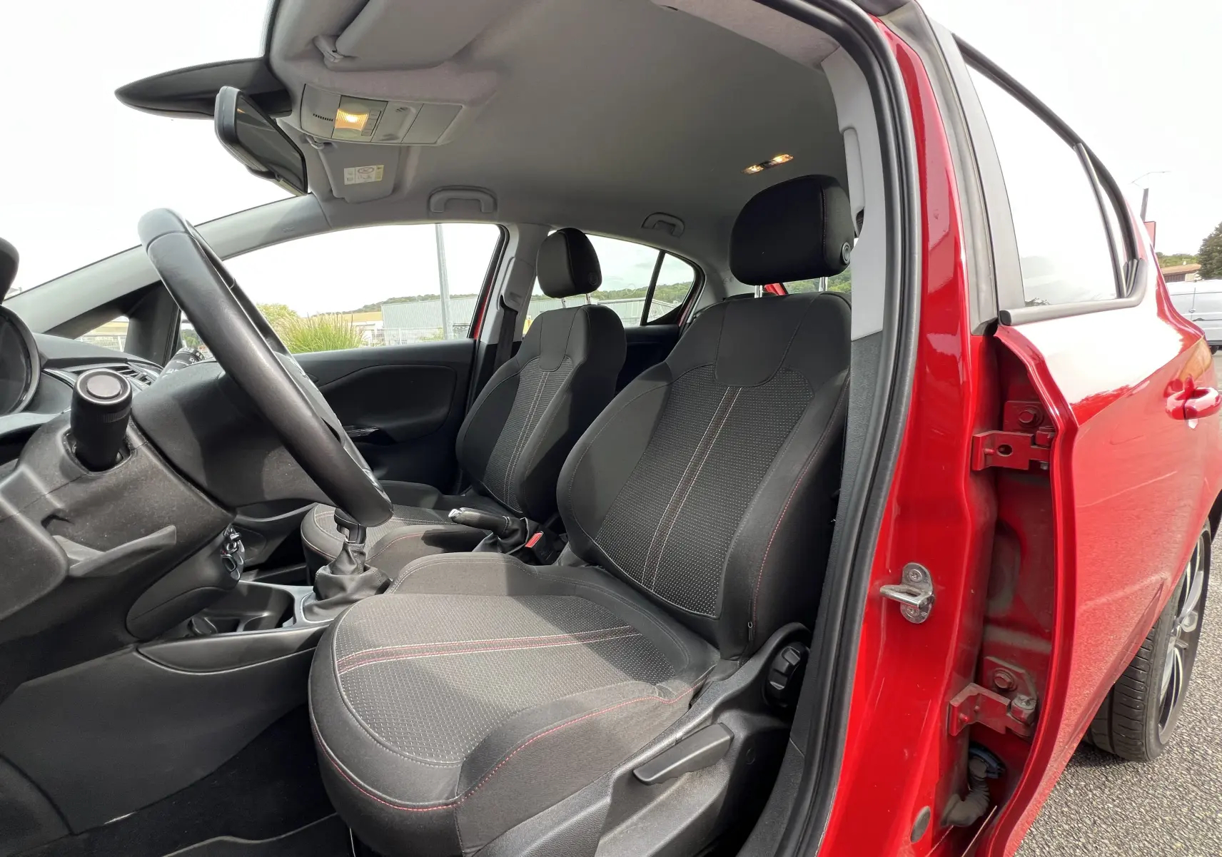 Vue intérieure côté conducteur d'une Opel Corsa rouge avec sièges tissu noirs à surpiqûres rouges et volant cuir.
