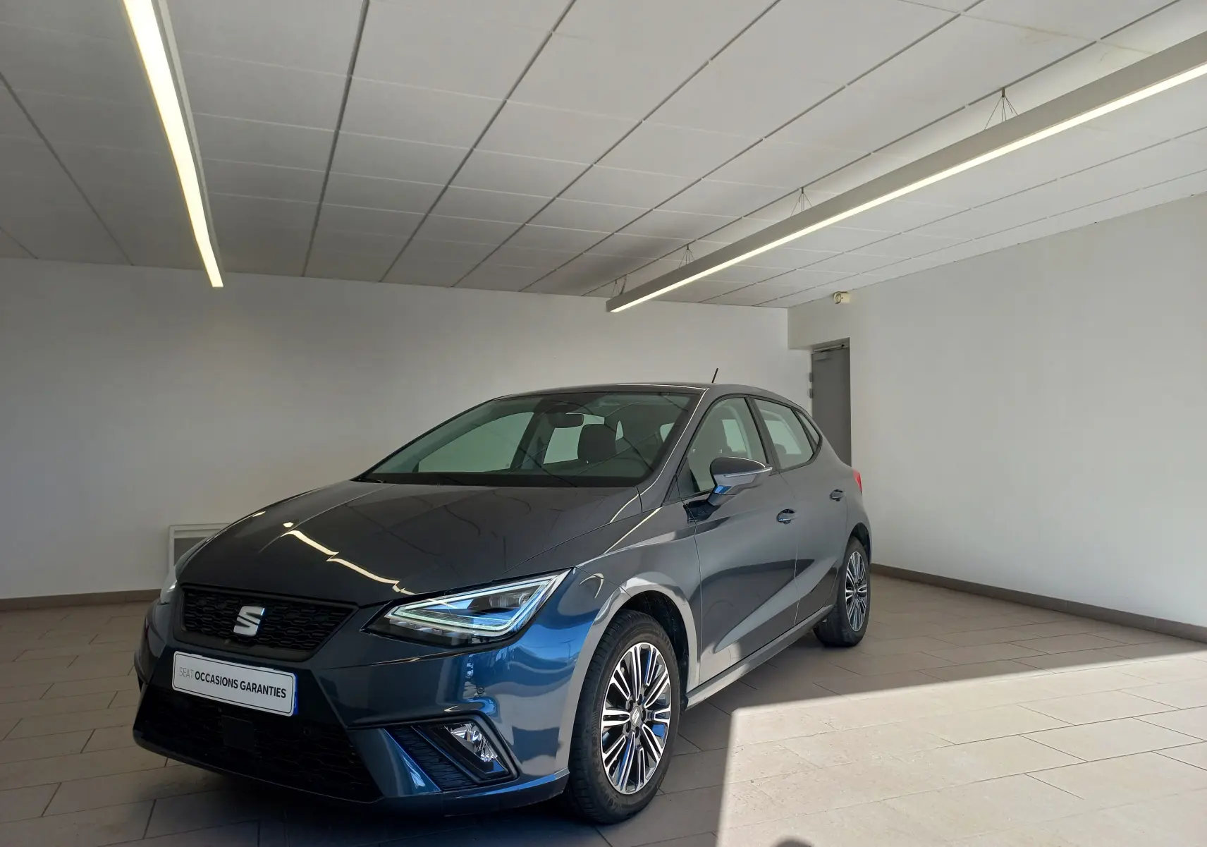 SEAT Ibiza 2025 gris magnétique vue 3/4 avant droit en intérieur avec jantes alliage et phares LED allumés.