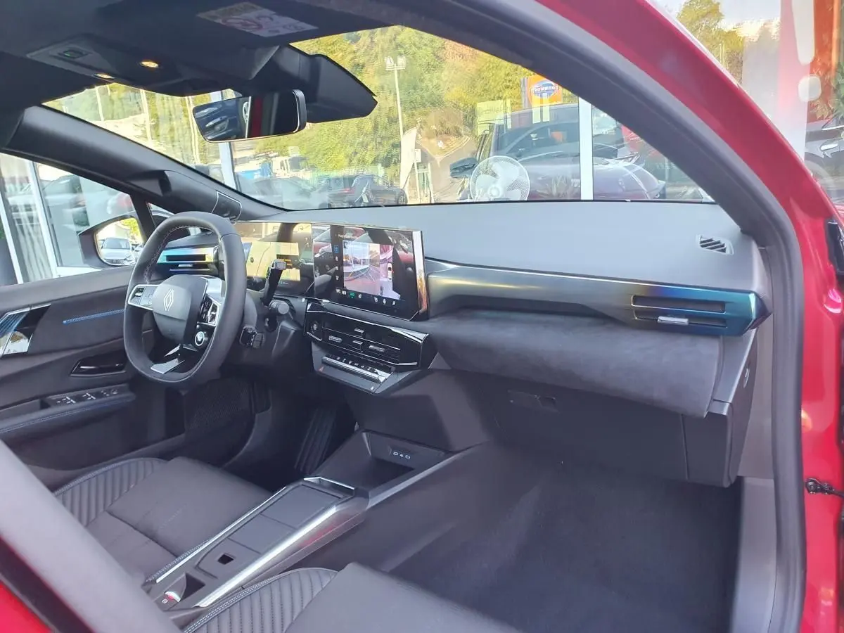 Vue intérieure côté conducteur de la Renault Clio VI rouge Absolu, avec tableau de bord moderne et écran tactile central.