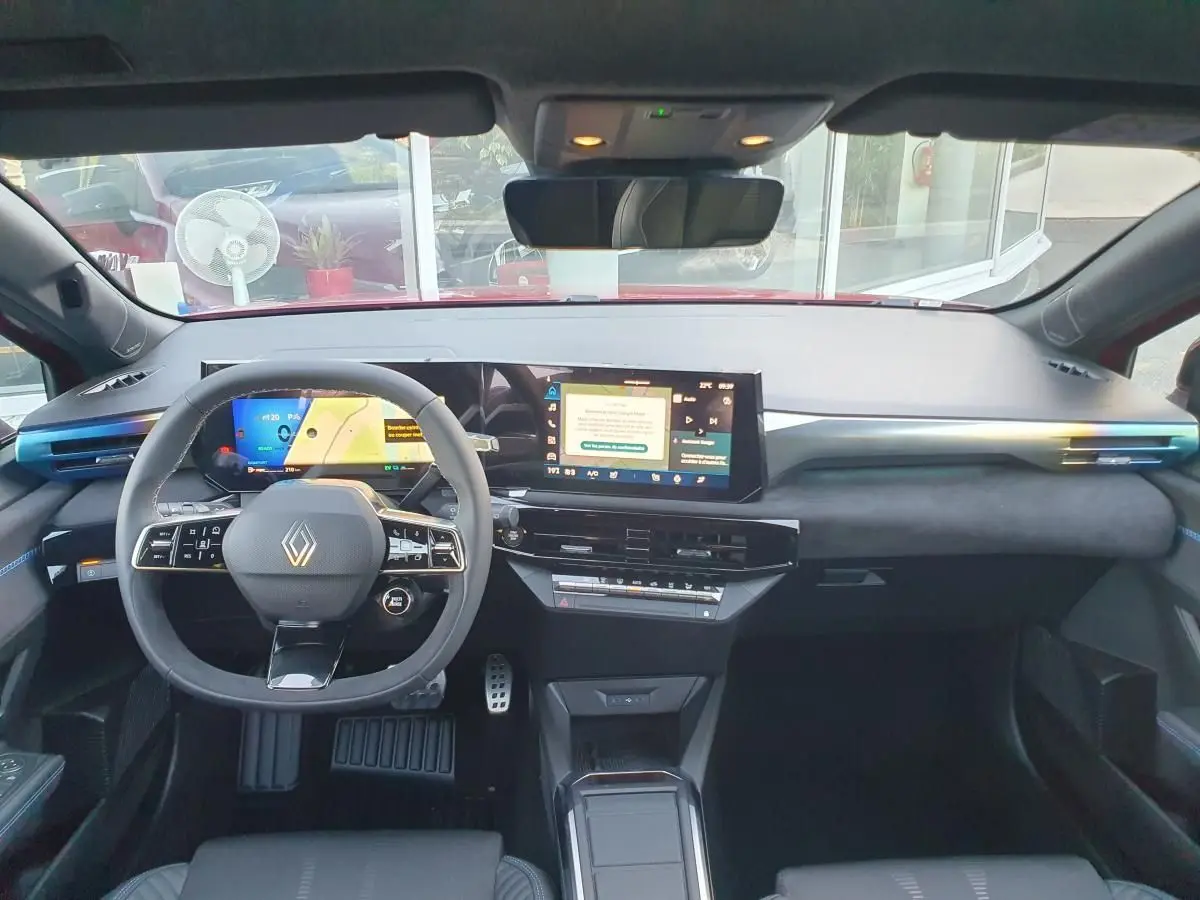 Vue intérieure avant de la Renault Clio VI hybride rouge, tableau de bord moderne avec double écran tactile et volant multifonction.