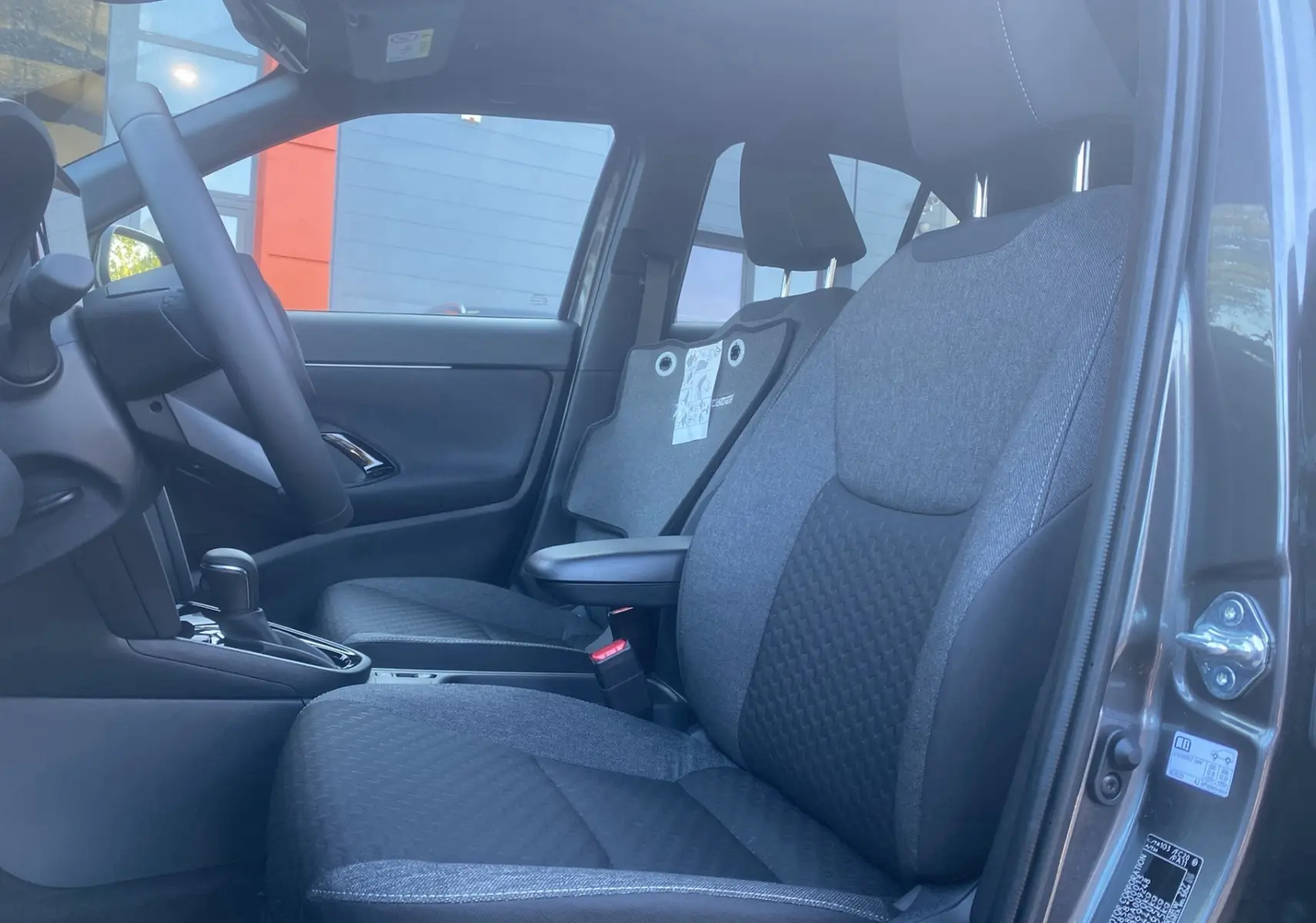Vue intérieure côté gauche des sièges avant gris tissu et volant cuir du Toyota Yaris Cross Hybride 130 Design Plus 2026.