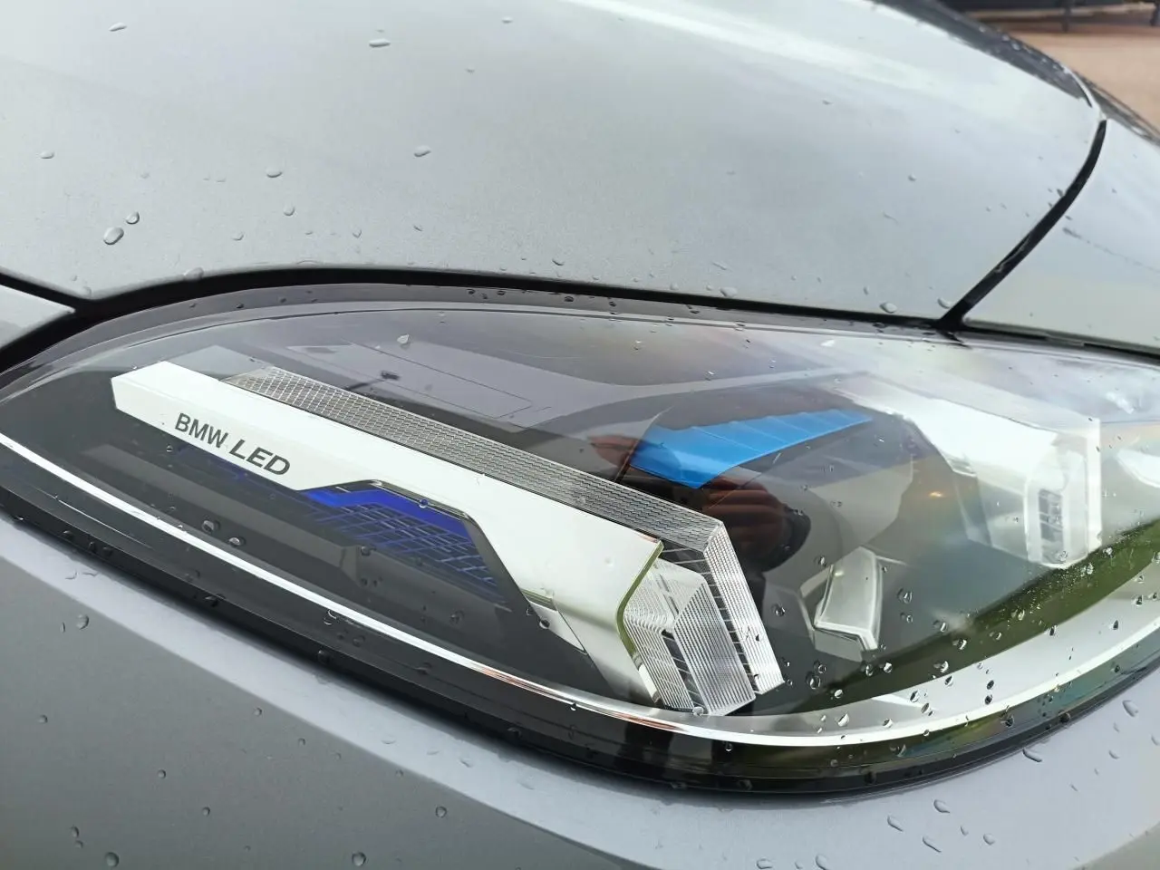 Gros plan sur le phare avant droit LED d'une BMW Série 1 gris Skyscraper avec toit noir, gouttes de pluie visibles.