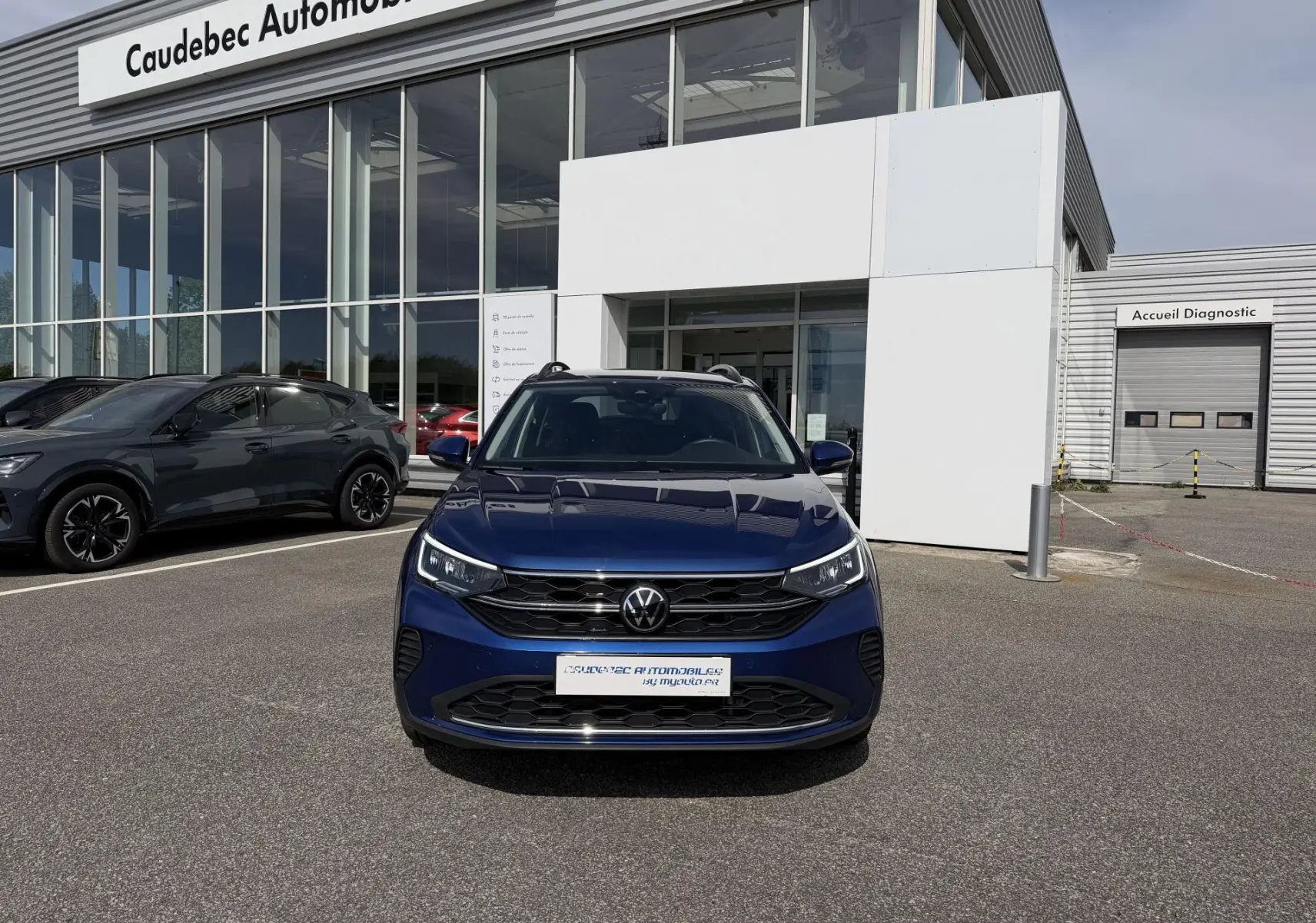 Vue de face du Volkswagen Taigo bleu métallisé garé devant un showroom Caudebec Automobiles par temps clair.