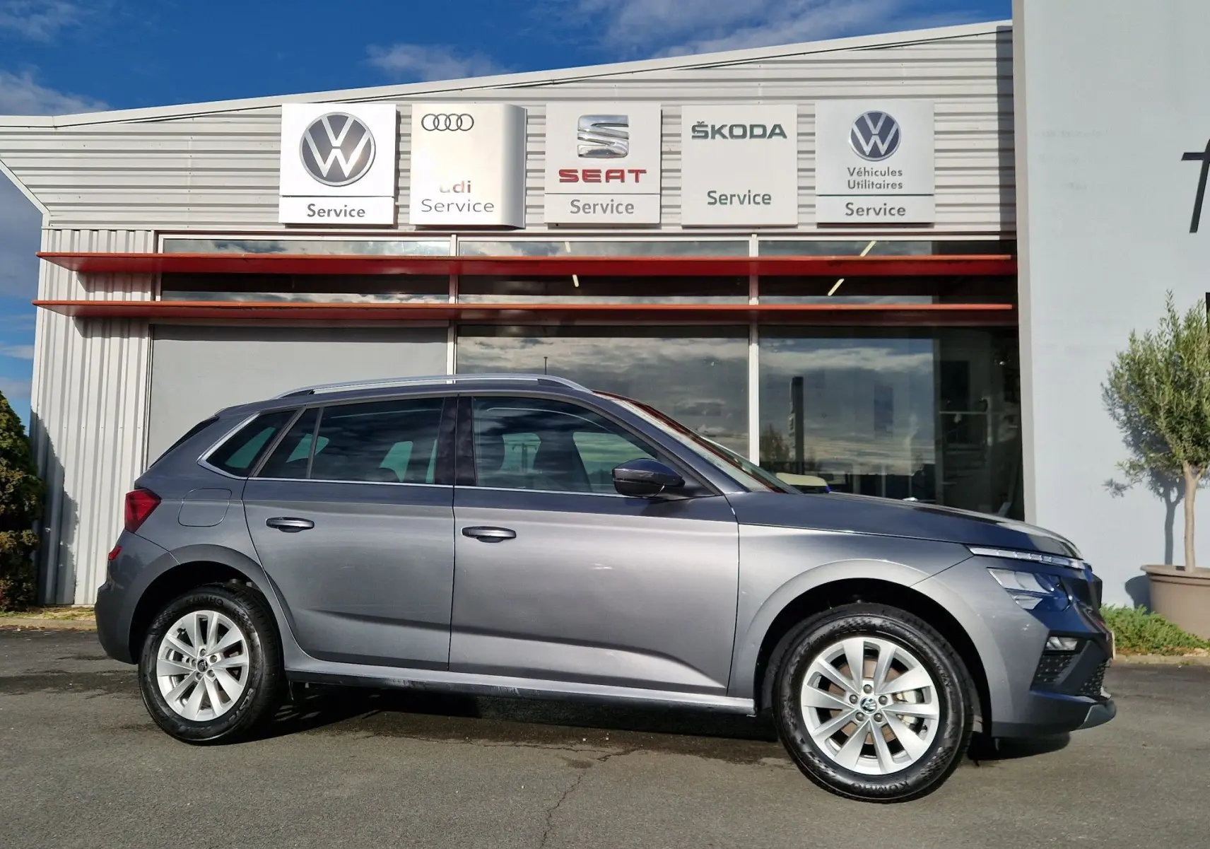 Vue de profil côté gauche d'un Skoda Kamiq gris graphite métal 2024 garé devant un service automobile.