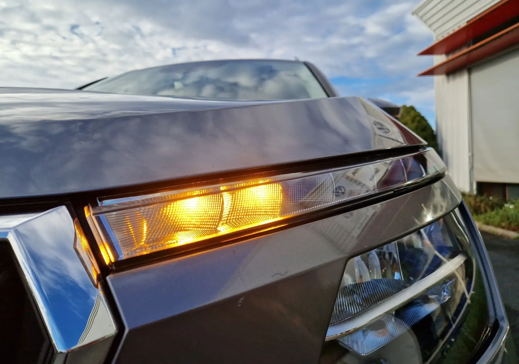Gros plan sur le feu avant droit allumé d'un Skoda Kamiq 2024 gris graphite métal sous un ciel partiellement nuageux.