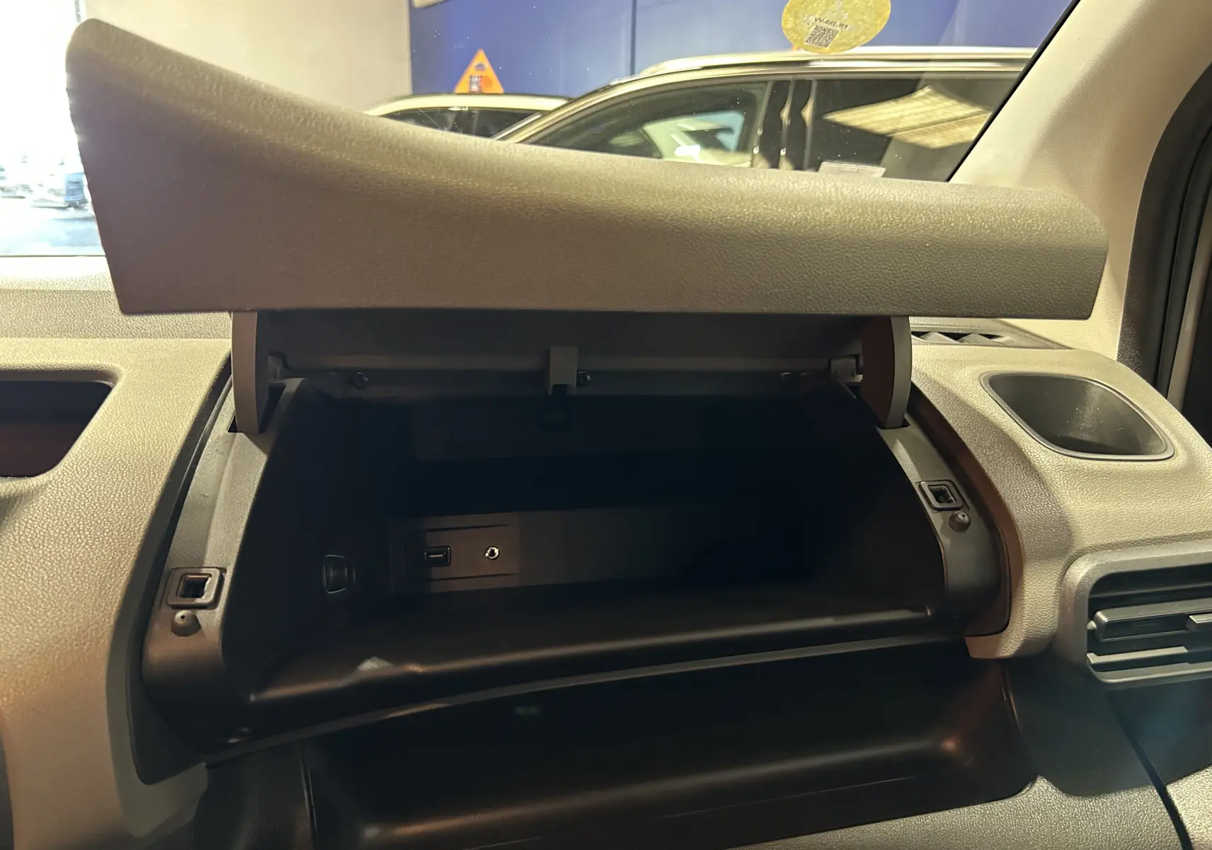 Gros plan sur la boîte à gants ouverte du Peugeot Rifter gris sable, montrant une prise USB et une prise auxiliaire.