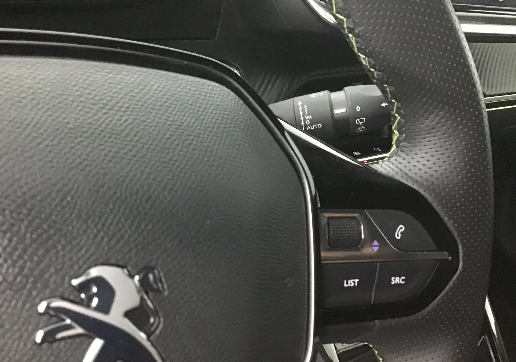 Gros plan sur le volant cuir perforé noir avec surpiqûres vertes du Peugeot 2008 électrique gris Artense, montrant les commandes audio.