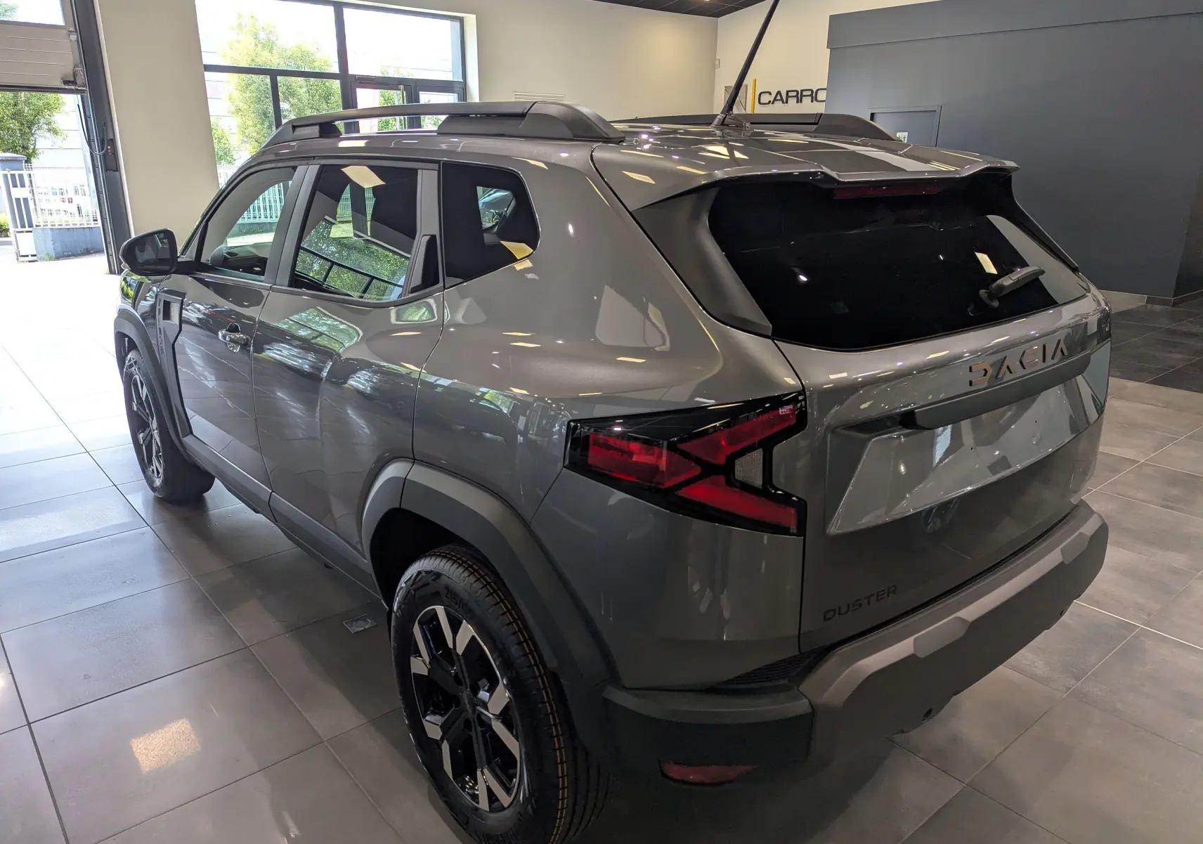 Vue 3/4 arrière droite du Dacia Duster 2025 gris schiste avec jantes noires et barres de toit en intérieur showroom.