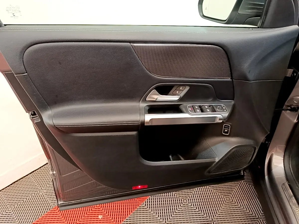 Vue intérieure côté gauche de la porte avant d’un Mercedes GLB 200 d Business Line gris foncé, avec commandes électriques et poignée argentée.