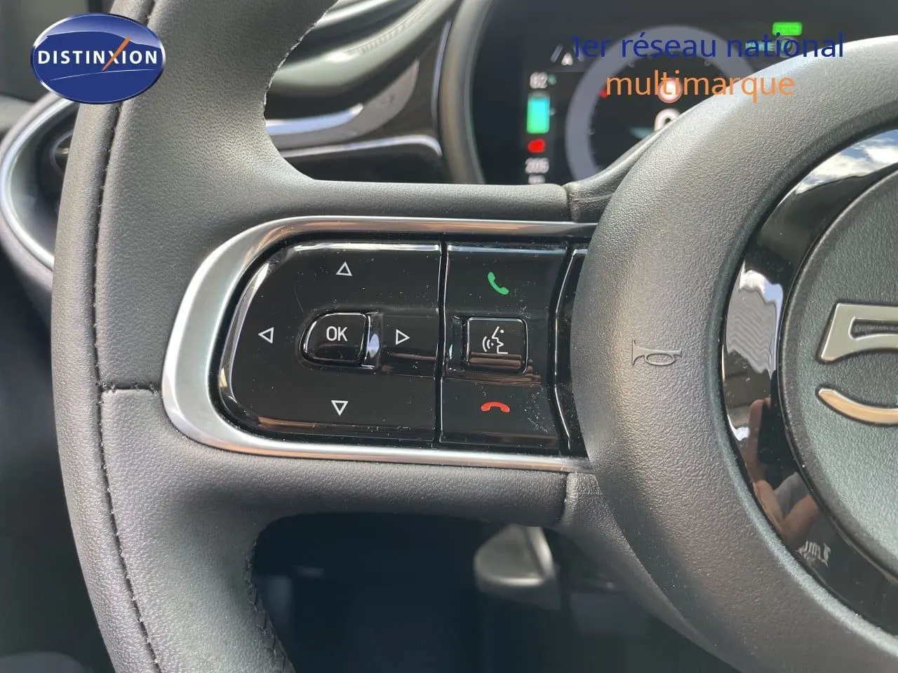 Gros plan sur le volant cuir noir de la Fiat 500 électrique 2023, avec commandes multifonctions et tableau de bord digital en arrière-plan.