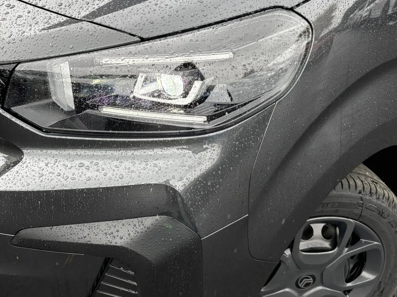 Gros plan sur le phare avant droit et la roue d'un Citroën Jumpy gris titane sous la pluie, avec détails LED visibles.