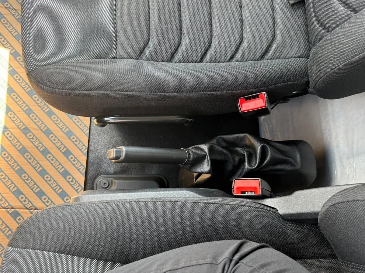 Vue rapprochée de la console centrale entre les sièges noirs du fourgon Iveco Daily, avec frein à main et boucles de ceinture rouges.