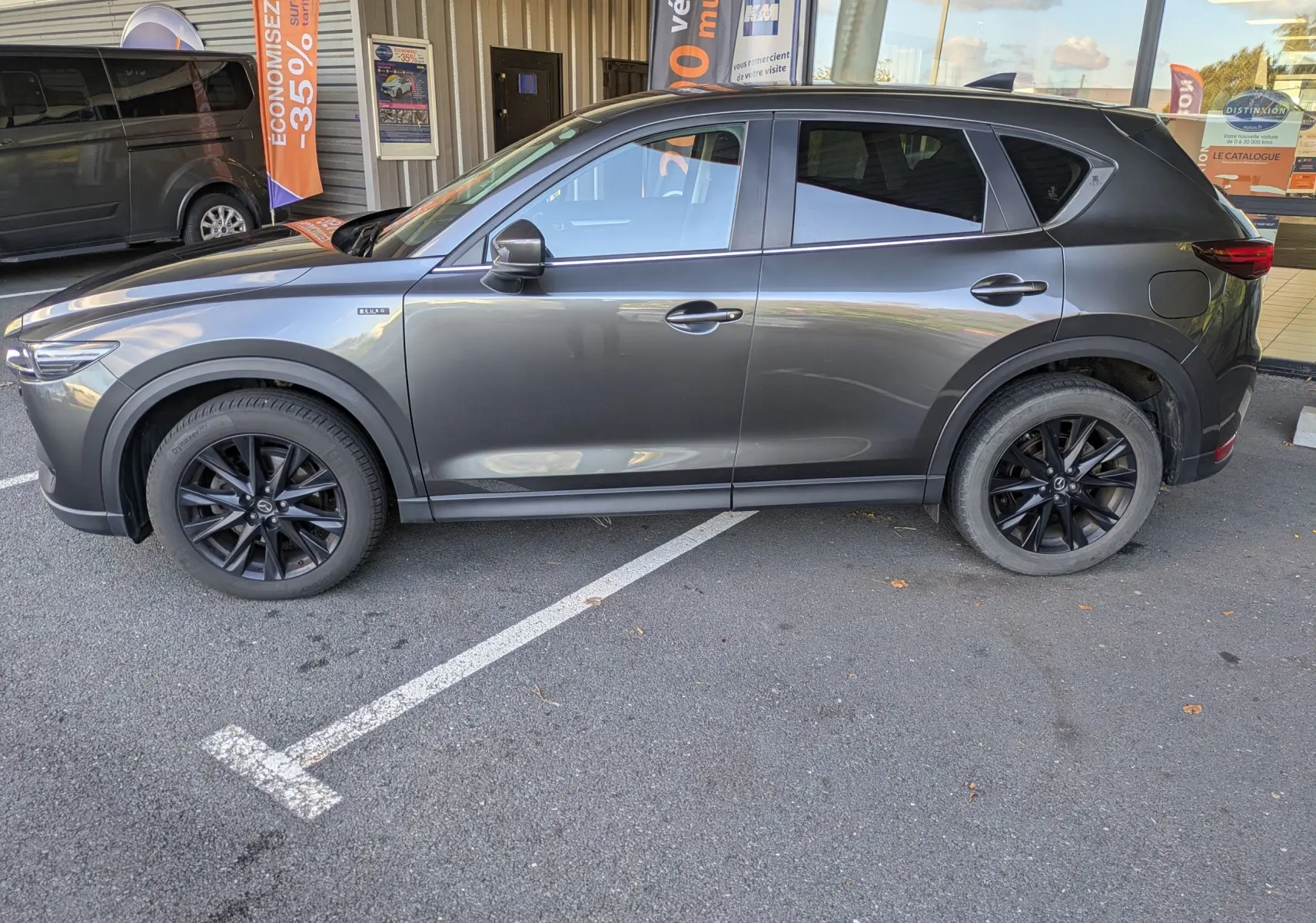 Mazda CX-5 gris 3/4 avant droit, version Kuro Edition avec jantes noires, stationné devant un bâtiment commercial.