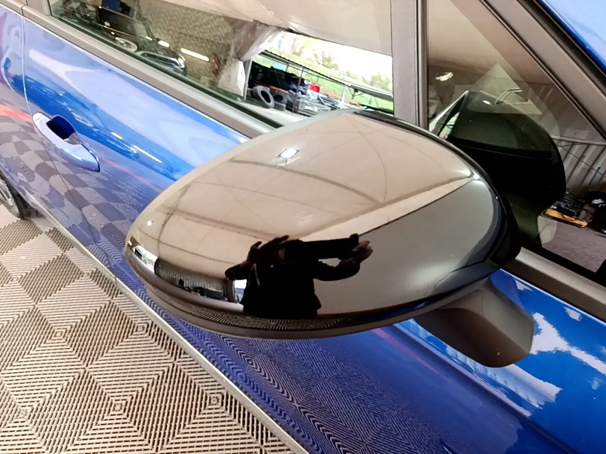 Gros plan sur le rétroviseur droit noir brillant de la Renault Clio Evolution bleu, avec reflet du plafond et de l'appareil photo.