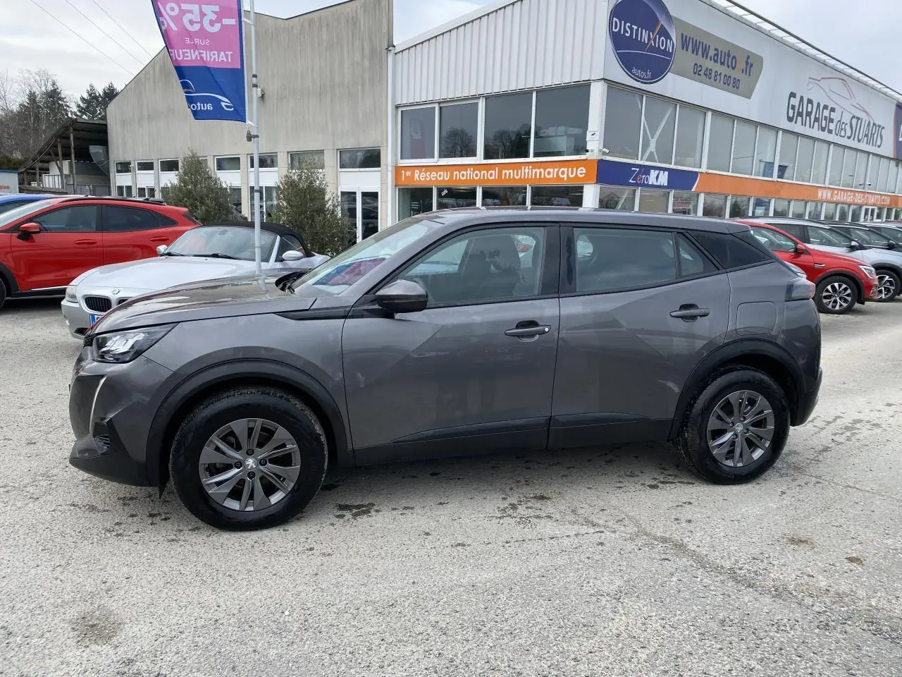 Peugeot 2008 gris foncé vue de profil côté gauche, stationnée devant un garage multimarque.