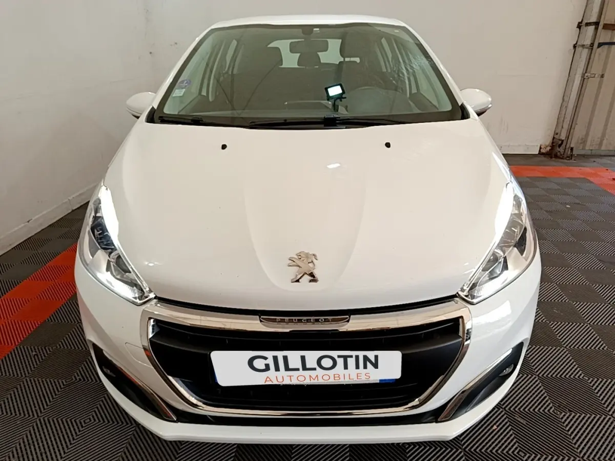 Vue avant d'une Peugeot 208 blanche avec calandre chromée et phares allumés dans un garage.