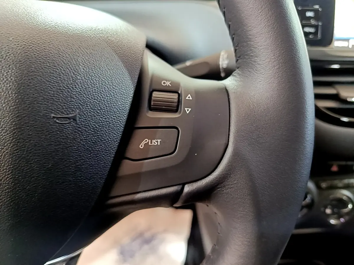 Gros plan sur le volant noir de la Peugeot 208 Puretech 82 S&S Active Business, avec commandes intégrées et tableau de bord flou en arrière-plan.