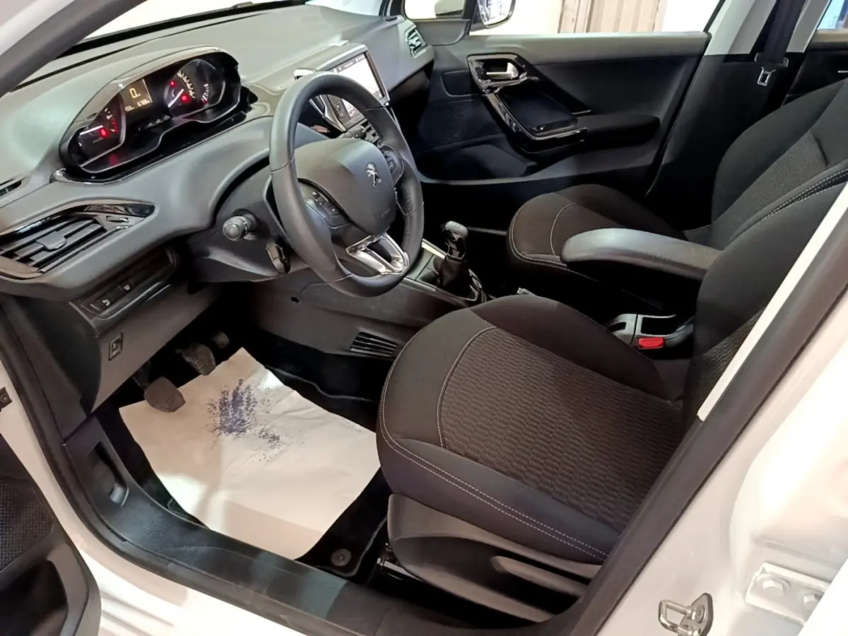 Vue intérieure côté conducteur de la Peugeot 208 blanche, montrant le tableau de bord et les sièges noirs.