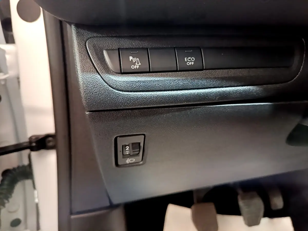 Détail du tableau de bord côté conducteur d'une Peugeot 208 blanche, avec boutons de contrôle et pédales visibles.