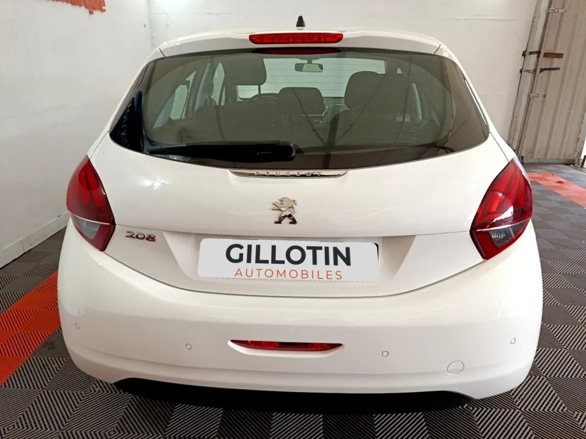 Vue arrière d'une Peugeot 208 blanche stationnée en intérieur, avec feux arrière rouges et logo lion central.